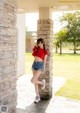 A woman leaning against a stone pillar in a red shirt and denim shorts.